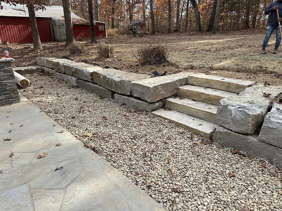 Stone retaining wall with steps in a gravel yard beside a concrete patio and wooded area