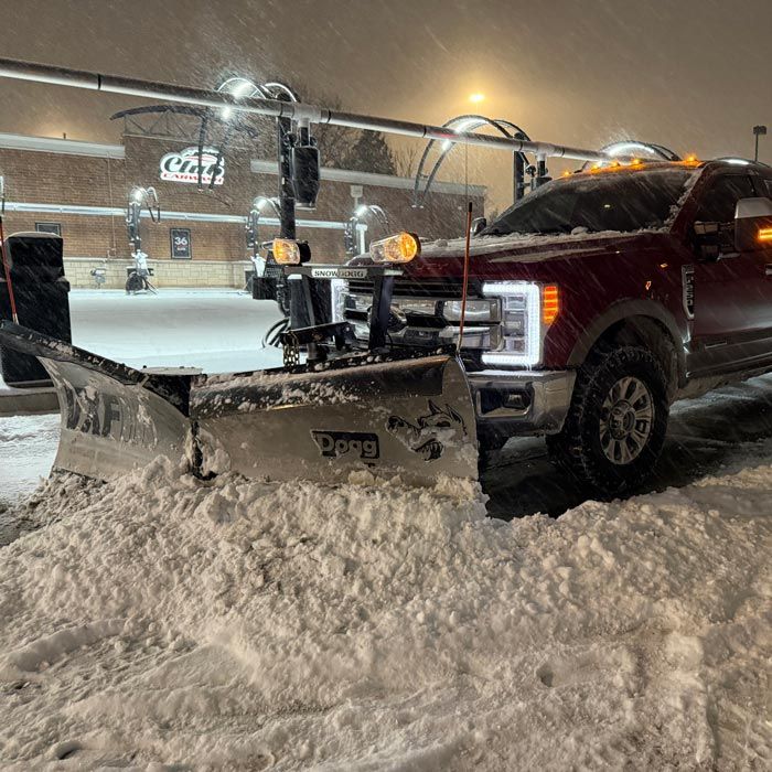 Red pickup with snow plow clearing a snowy parking lot at night