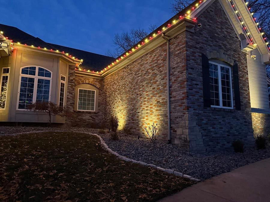 Stone house at dusk with warm holiday lights along the roofline and lit windows.