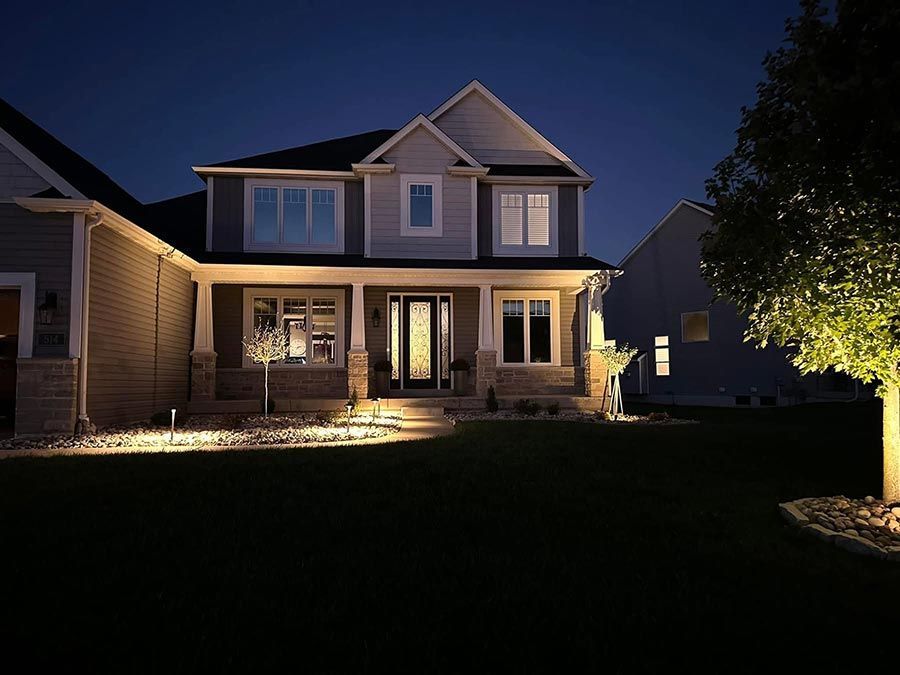 Two-story suburban house at night with warm porch lights and a lit front yard