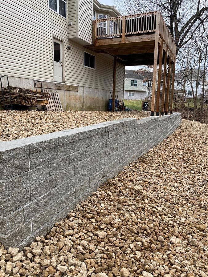 Stone retaining wall beside a house deck, with gravel yard and leaf-covered ground under bare trees.