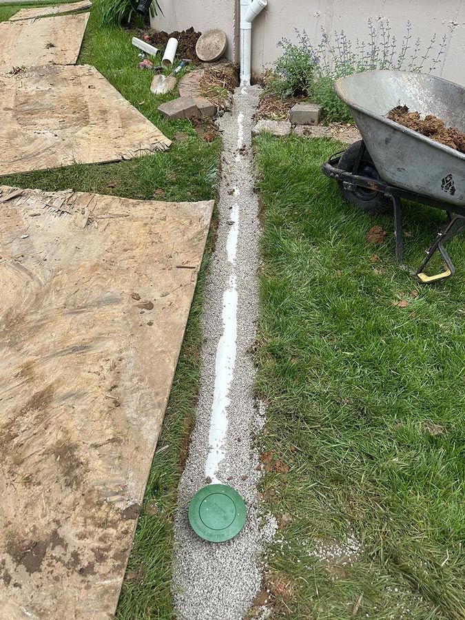 Narrow concrete drain channel with green cap beside grass, pavers, and a wheelbarrow in a yard.