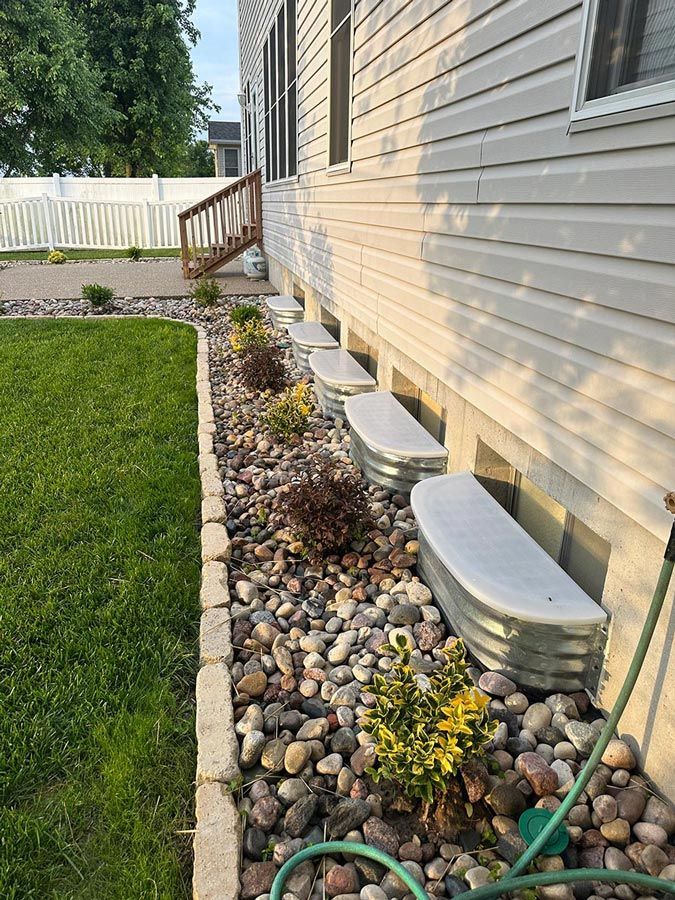 Side yard with river rocks, white stepping stones, garden hose, and beige house siding beside a lawn.