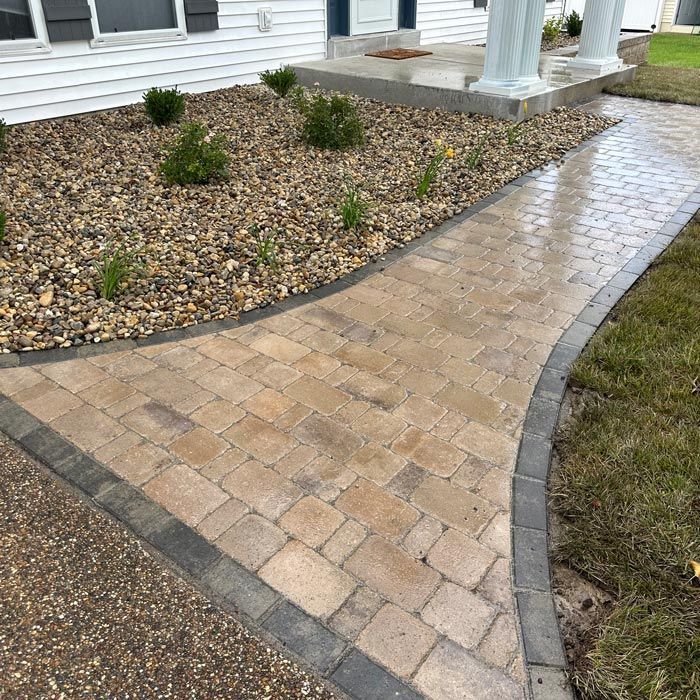 Curved brick walkway beside a white house and porch, bordered by gray pavers and gravel landscaping.