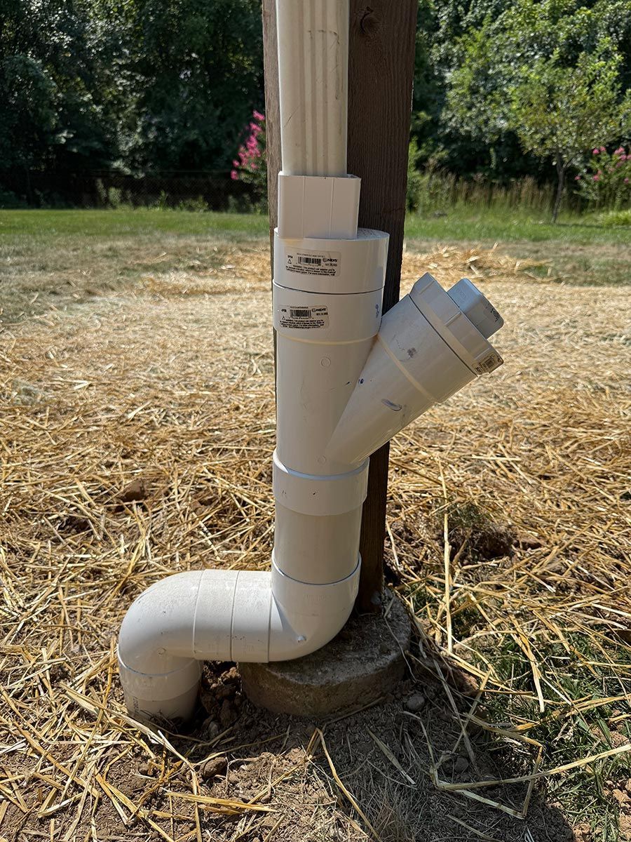 White PVC downspout drainage setup beside a wooden post on straw-covered ground outdoors