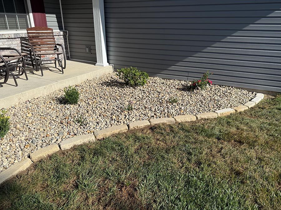 Rock garden bed with small shrubs along a house exterior, bordered by stone and lawn.