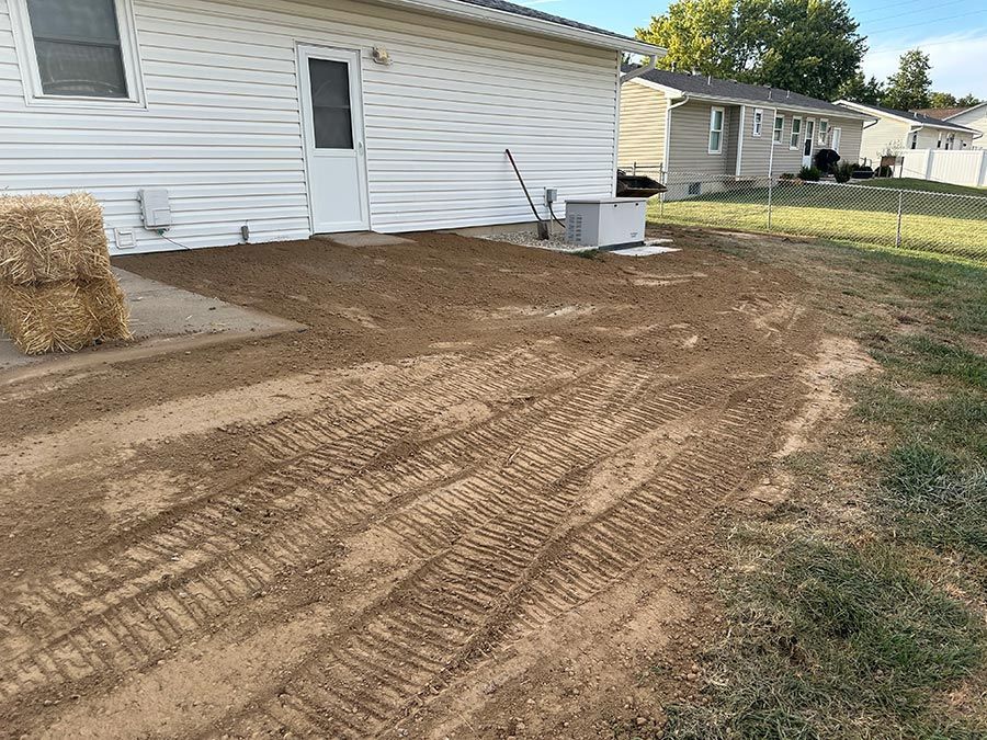 Freshly graded dirt yard beside a white house, with hay bales and a small utility box near the wall.