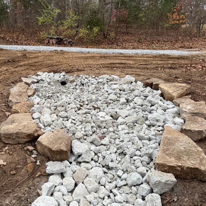Rock-lined drainage swale filled with white gravel beside a dirt road in a wooded area