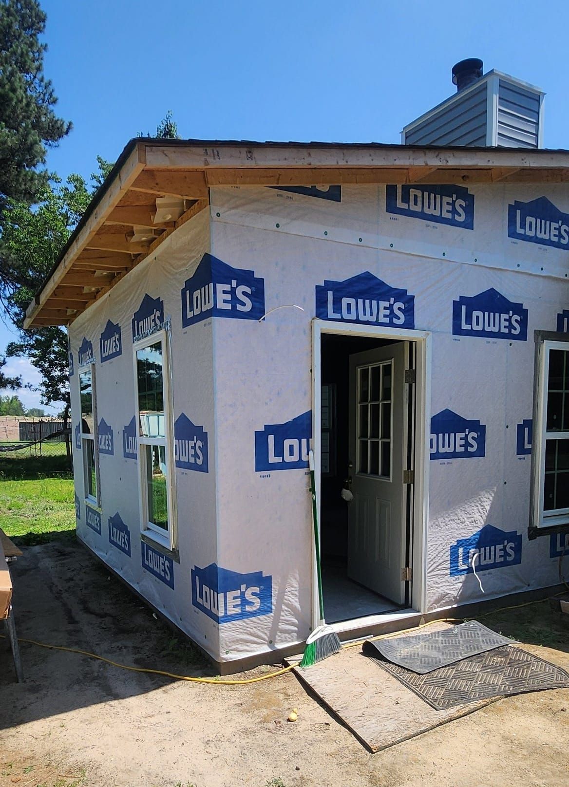 Exterior view of a small building under construction, with Lowe's wrap, windows, door, and exposed roof framing.