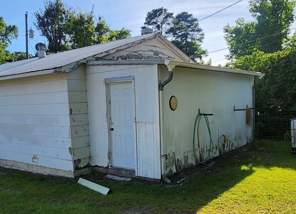 White shed with peeling paint, door, and green hose on the side, set in a grassy yard.