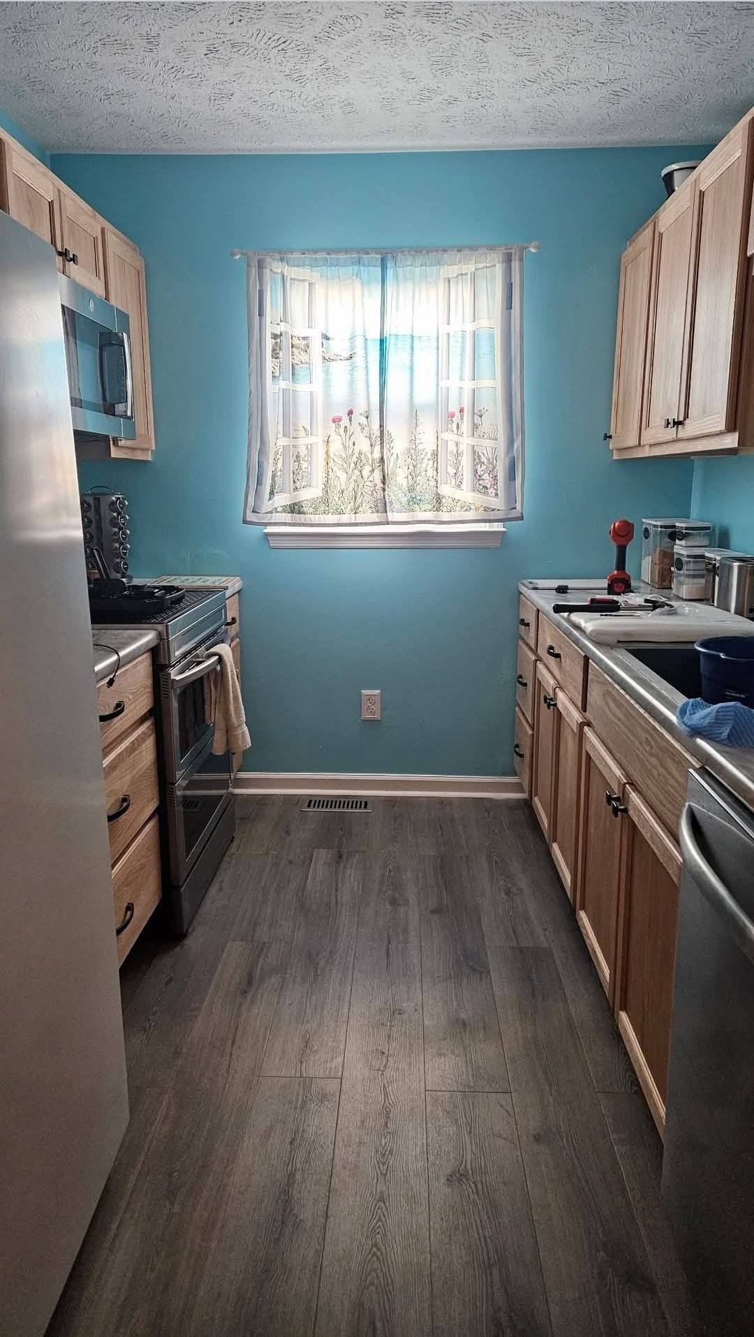 Narrow kitchen with wood cabinets, light blue walls, and a window. Gray flooring.