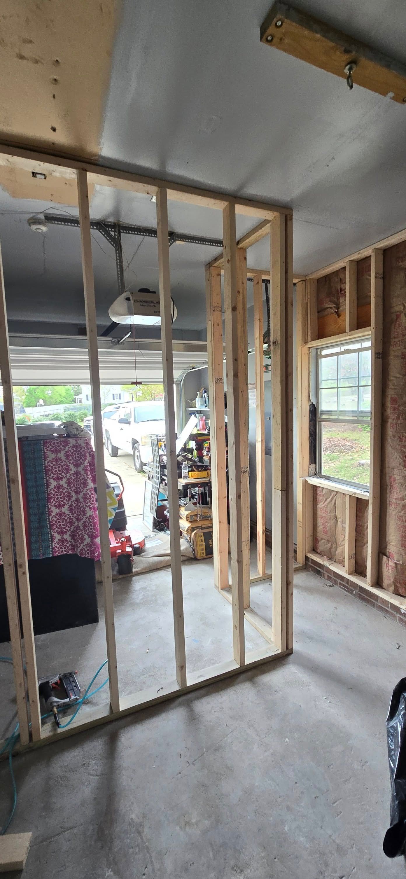 Wooden studs form interior walls and ceiling in a garage, with a window and the open garage door visible.