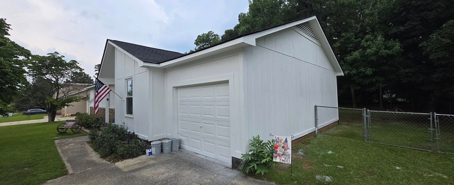 White house with black roof, garage door, American flag. Green grass, trees. Cloudy sky.