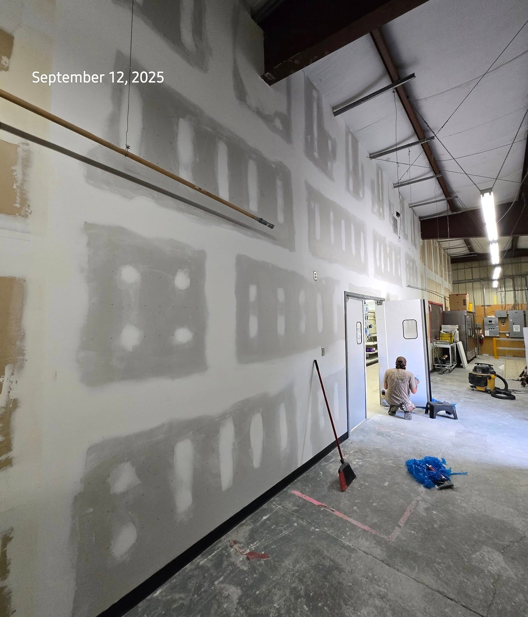 Interior view of a construction site: drywall installation on a wall, worker in the background, tools on the floor.