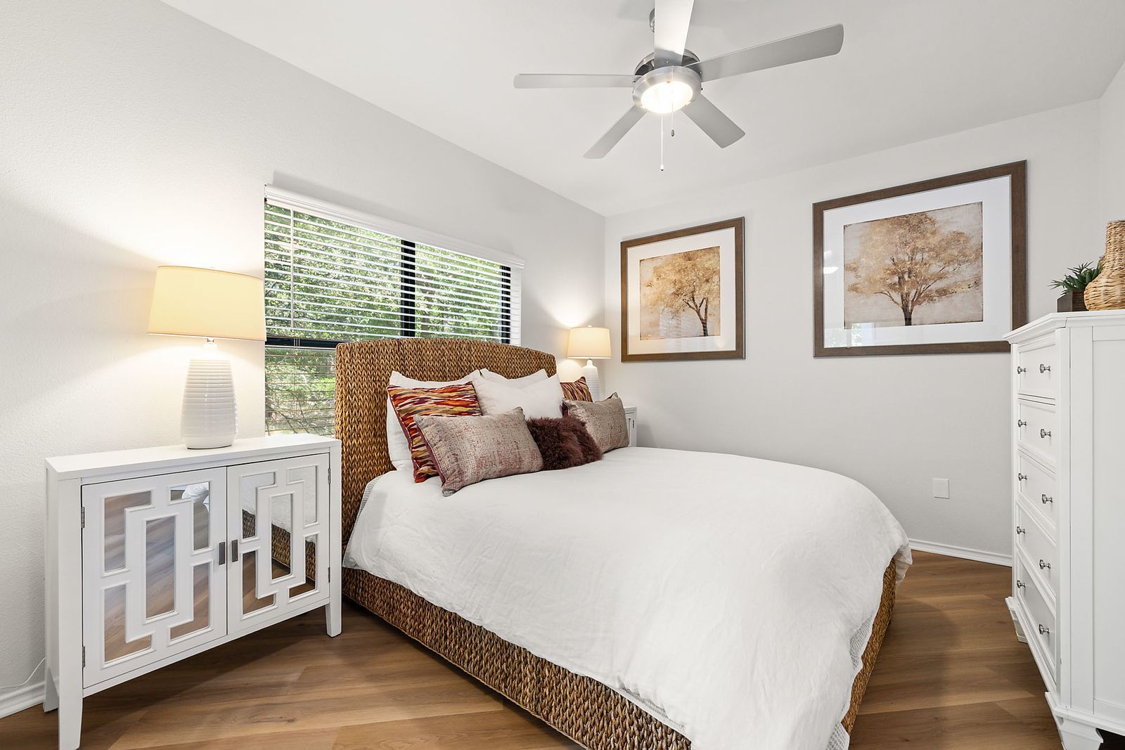 Bright bedroom with white bedding, wood floor, decorative pillows, framed art, and a ceiling fan.