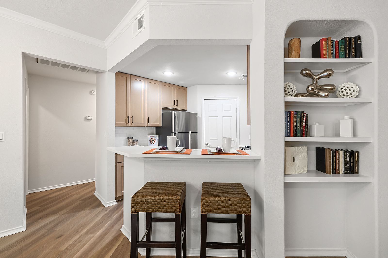 Bright kitchen view with white walls, wood floors, bar stools, and built-in shelves with books and decor at Marquis at Caprock Canyon in Austin, TX.