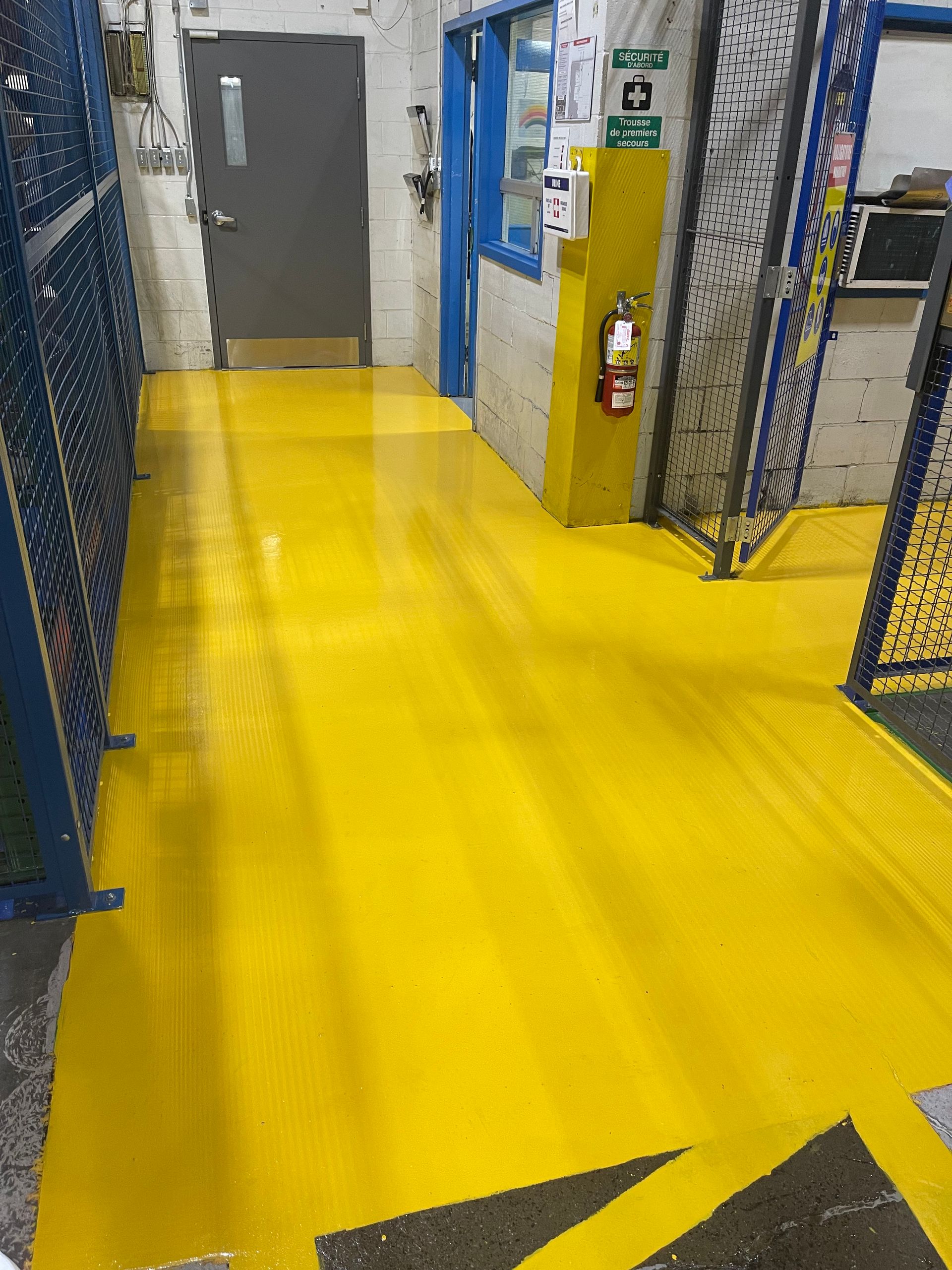 Sol peint en jaune dans un environnement industriel avec cages de sécurité bleues, porte et extincteur.