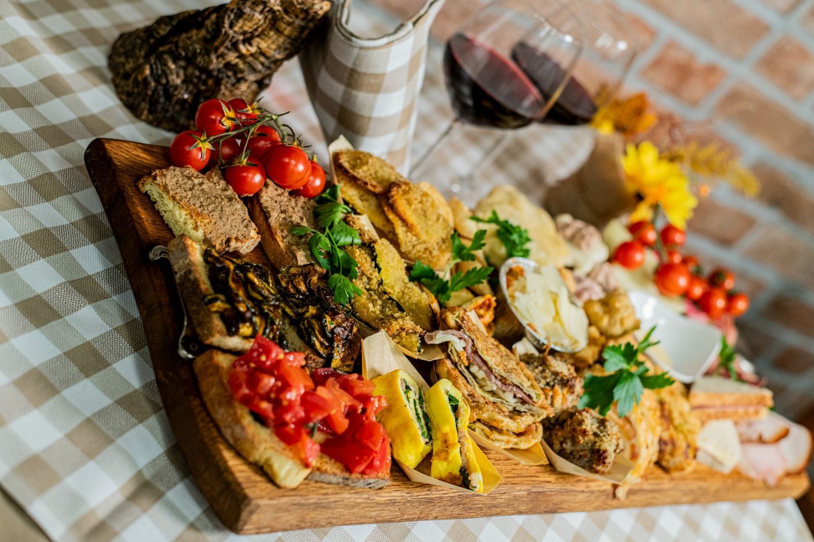 Un tagliere di legno con un assortimento di carni grigliate, formaggi, erbe aromatiche e pomodorini, accanto a bicchieri da vino su una tovaglia a quadri.