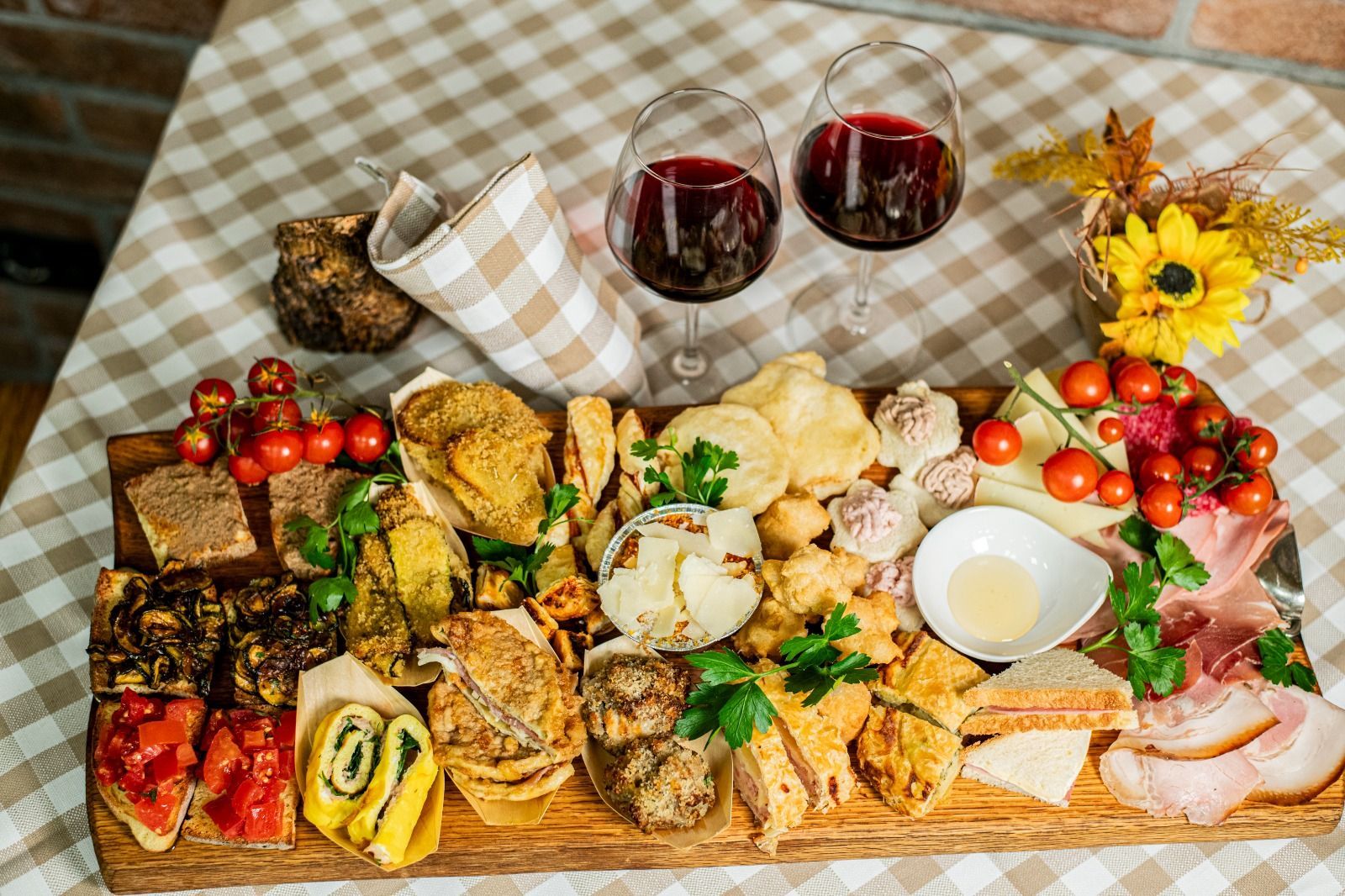 Un assortimento di pasticcini, snack e frutta disposti su una tovaglia a quadri, accompagnati da due bicchieri di vino rosso.