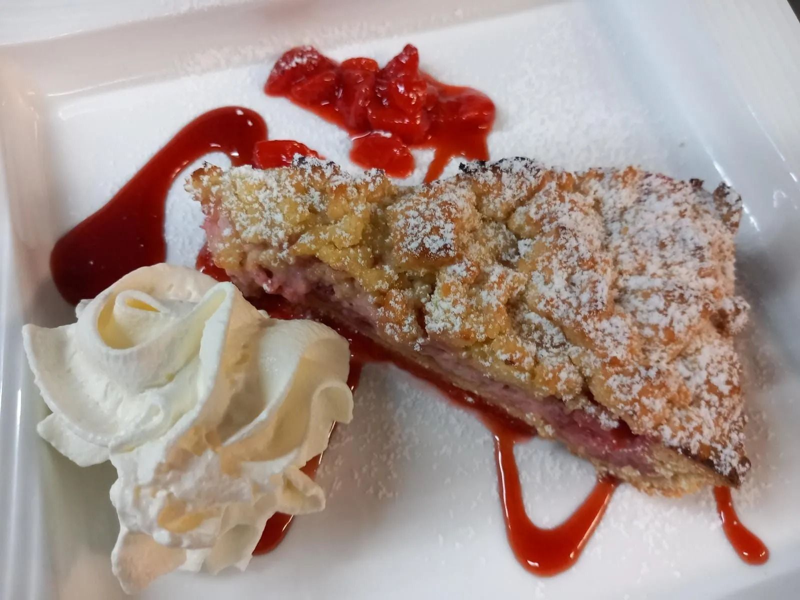 Fetta di crostata di frutti di bosco con zucchero a velo, panna montata e salsa di fragole su un piatto bianco.