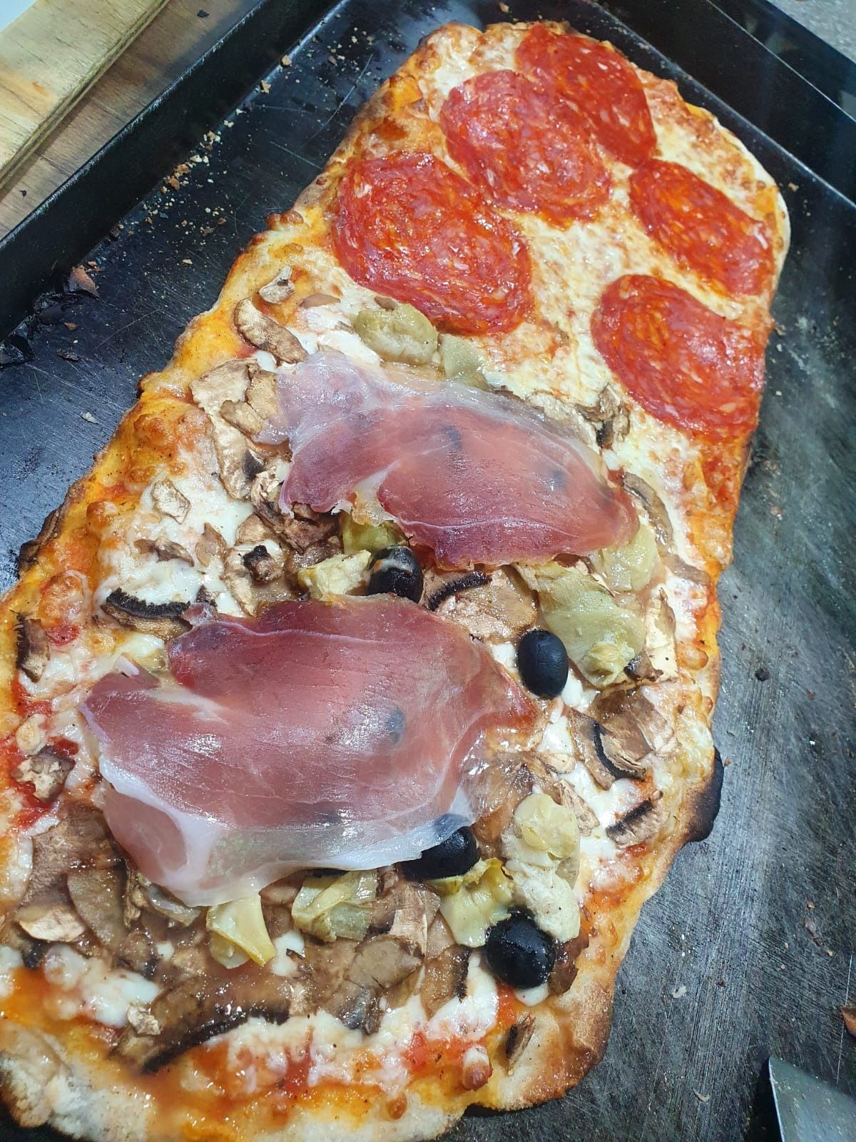 Pizza rettangolare su una teglia da forno, guarnita con peperoni, fette di prosciutto e funghi.