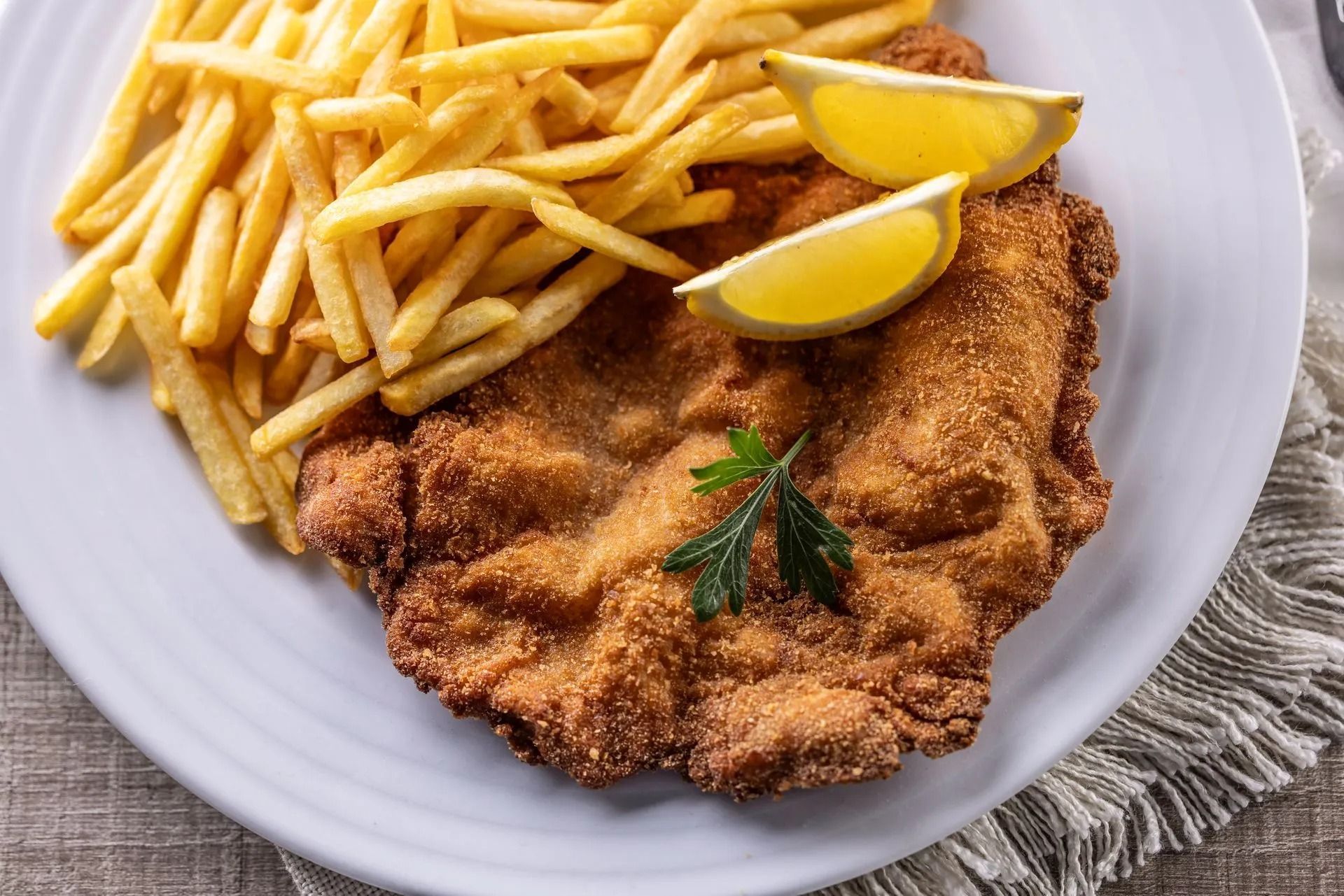 Cotoletta impanata con patatine fritte e spicchi di limone su un piatto bianco.