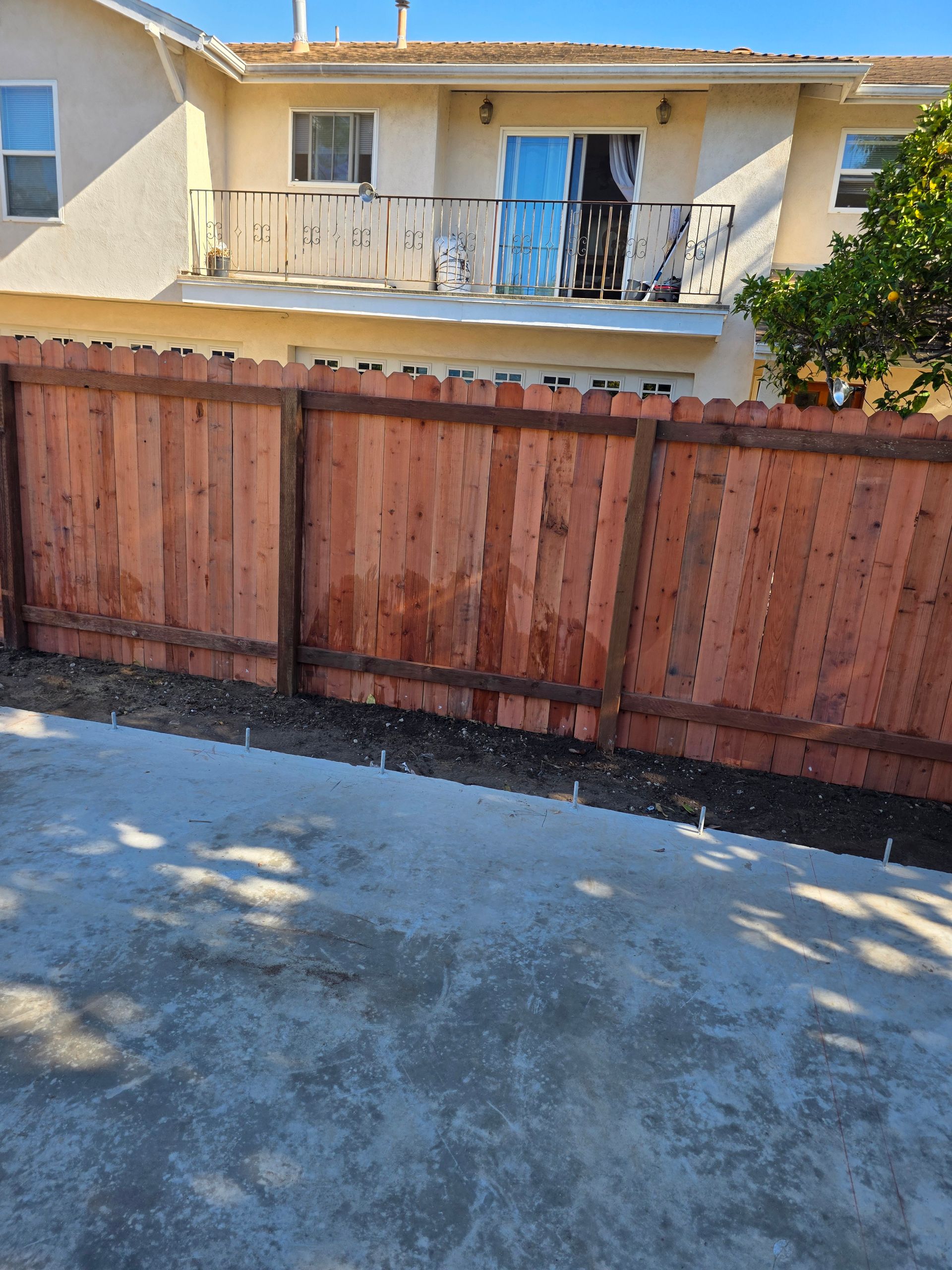 Wooden fence in front of a beige apartment building with balconies and a small tree.