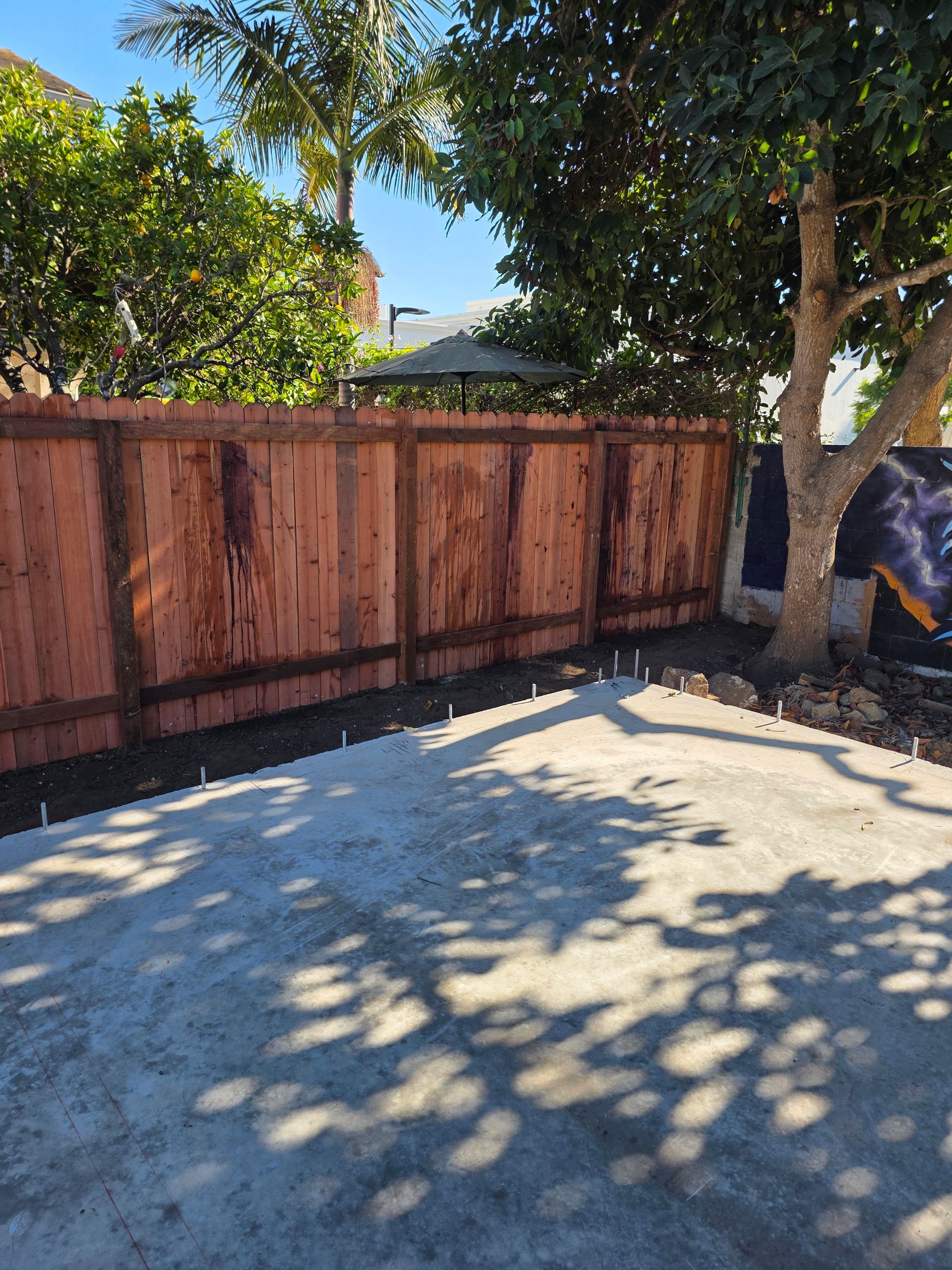 Wooden fence along a sunlit driveway with trees casting shadows and a person working near the right edge