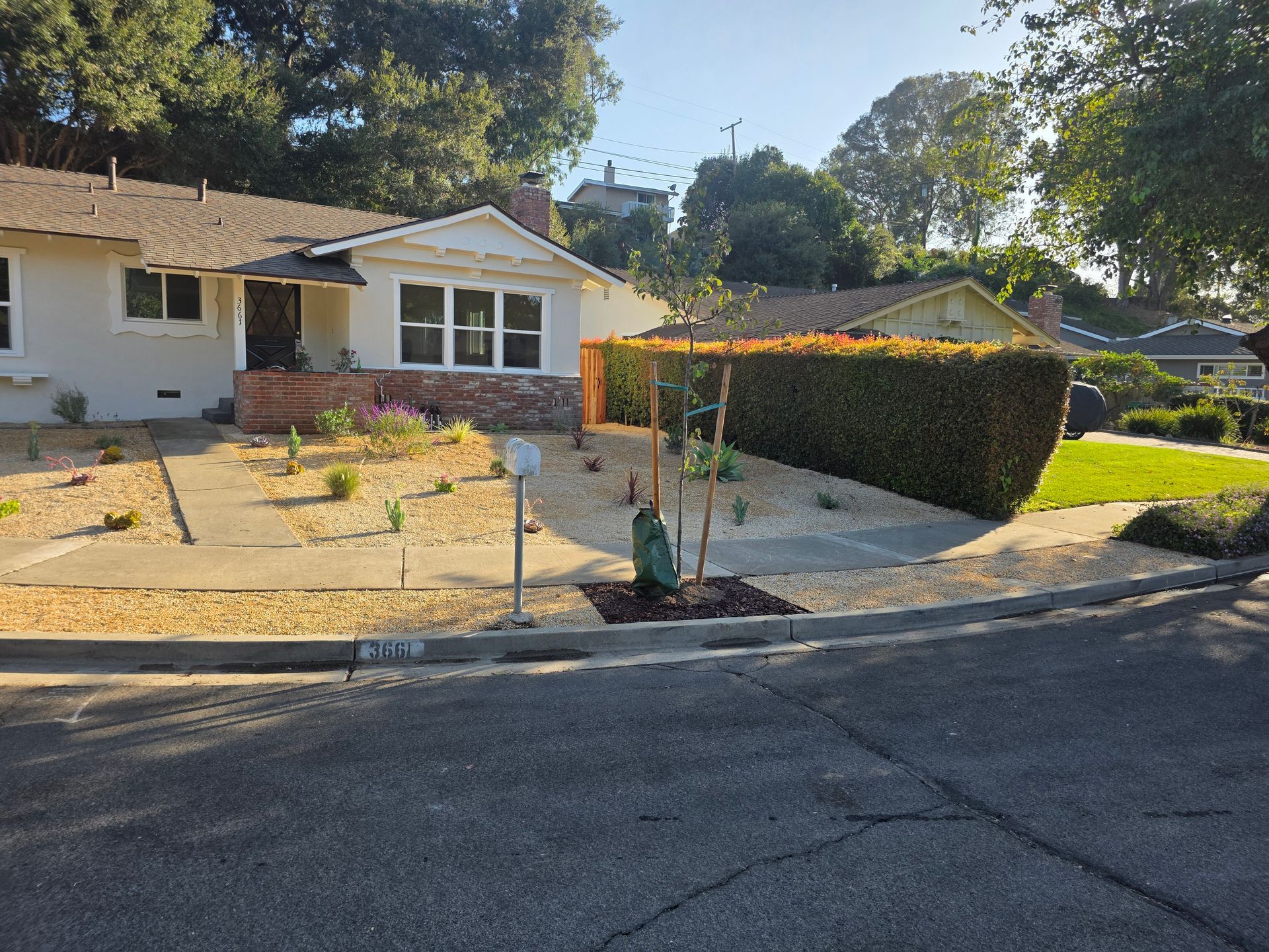 Single-story suburban house with a gravel front yard, shrubs, and a trimmed hedge by the street.