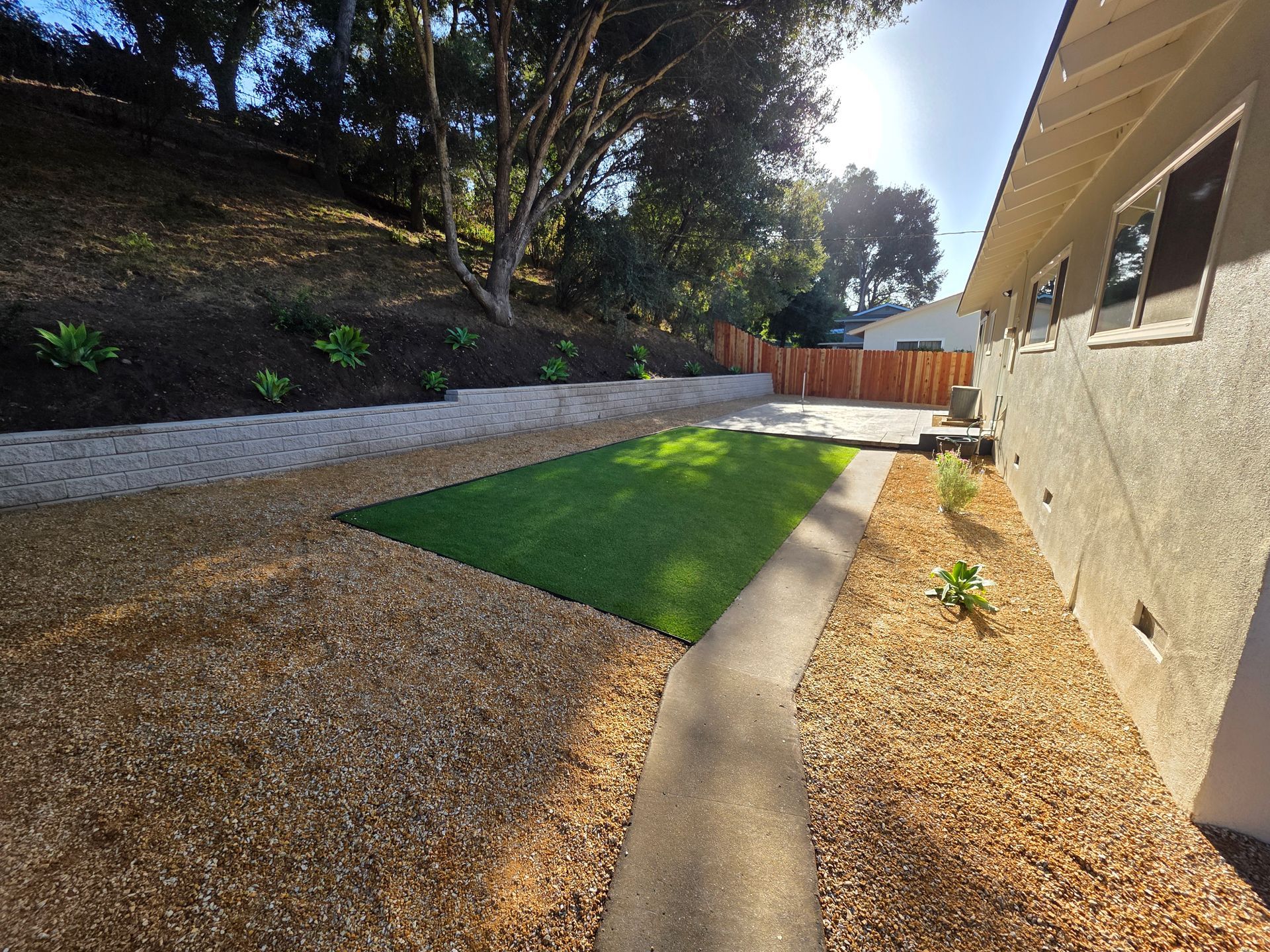 Backyard with gravel path, green turf strip, retaining wall, and beige house in sunlight