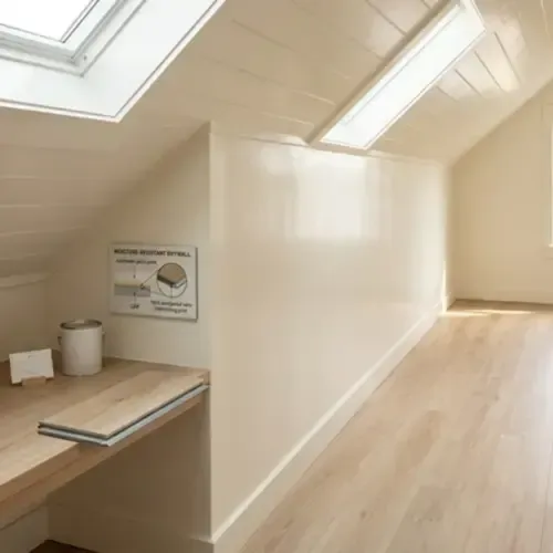 A bright attic room with light wood flooring, a sloping white ceiling, skylights, and a small built-in wooden desk area.