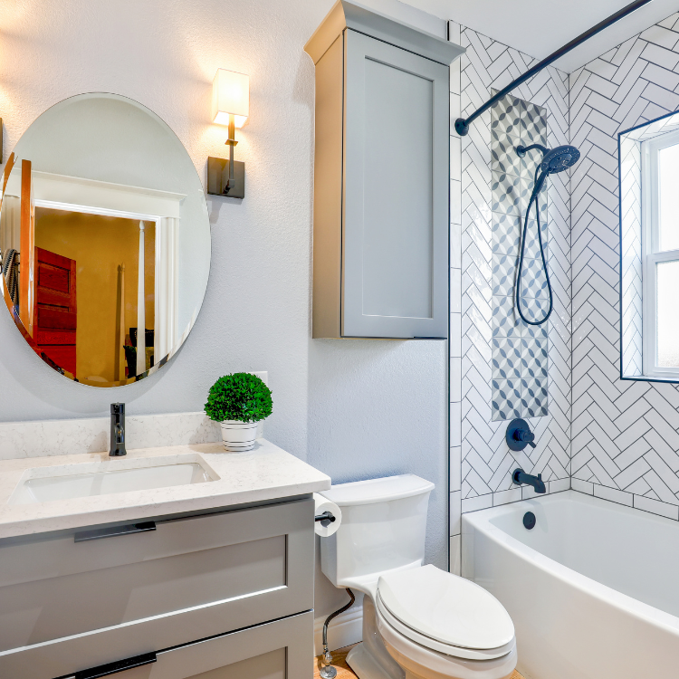 A modern bathroom with a gray vanity, oval mirror, white toilet, and a bathtub with white herringbone tiled walls.