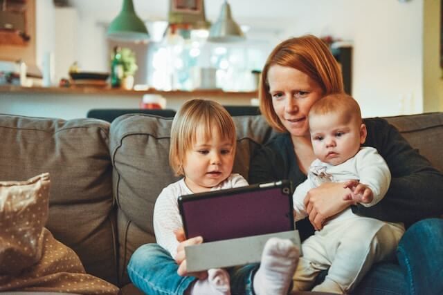 Woman on a couch with two small children, one holding a tablet in a cozy living room.