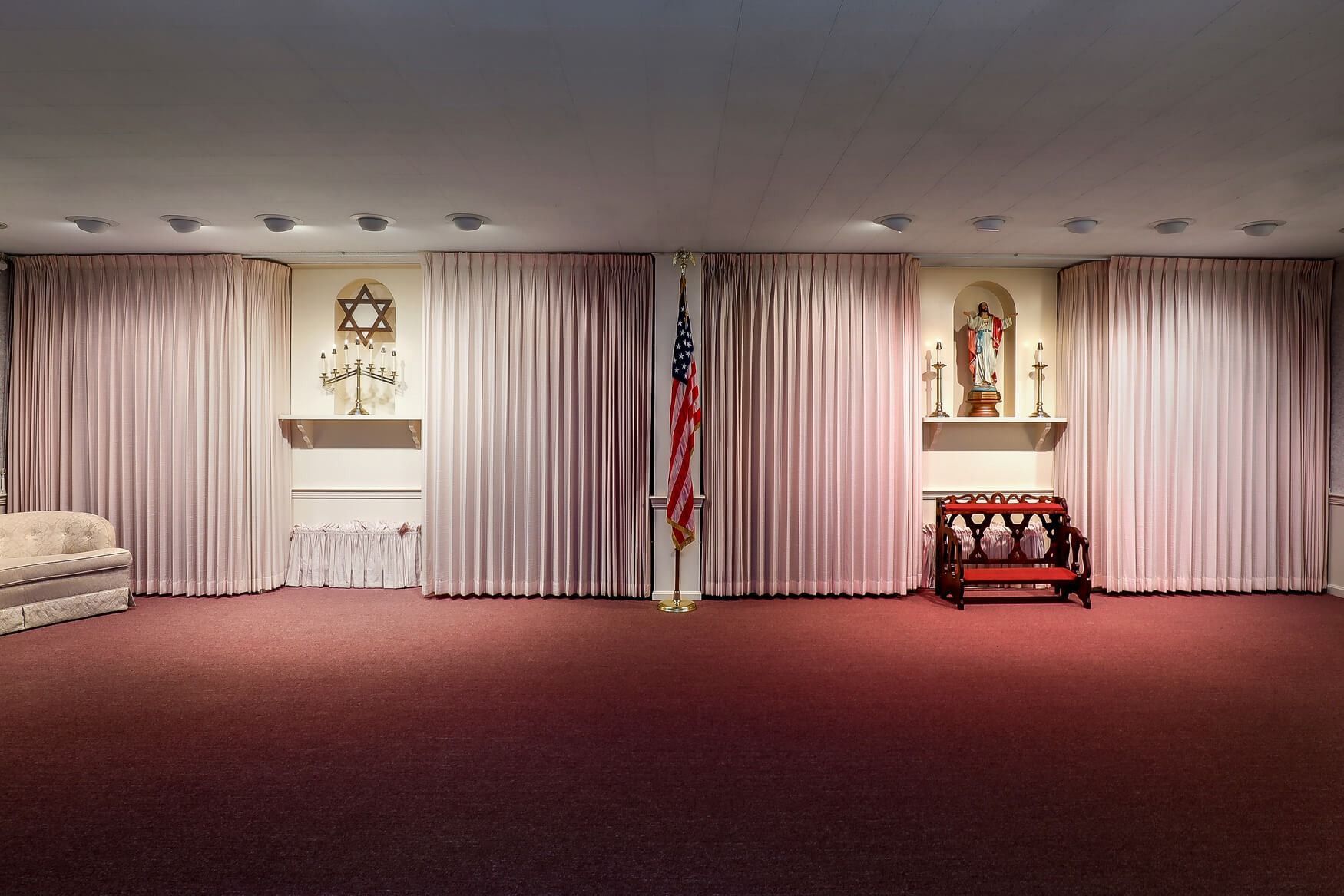 Empty ceremonial room with red carpet, pink curtains, U.S. flag, and a small religious altar at center-right