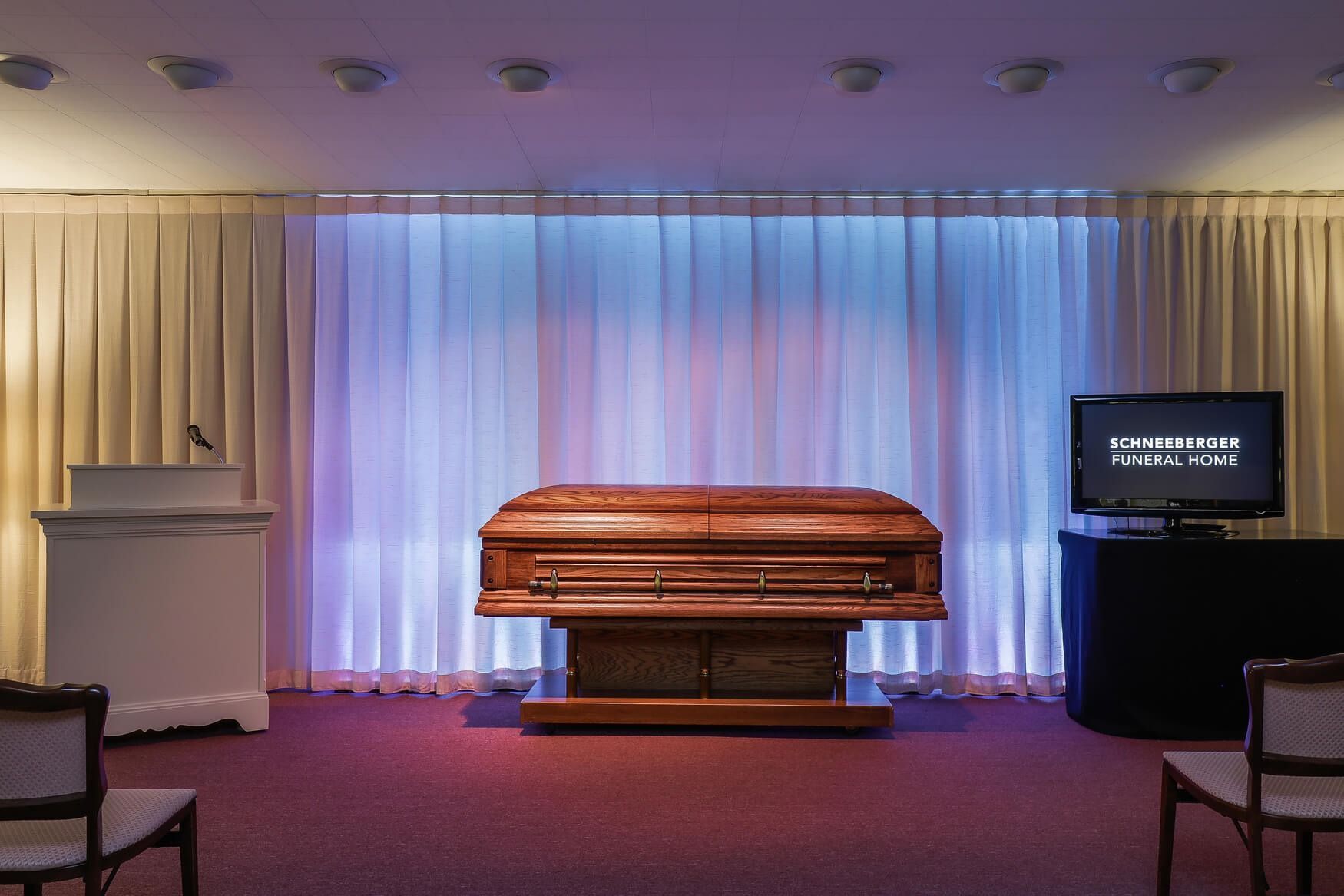 Wooden casket on a stand in a funeral chapel with white drapes and soft blue lighting