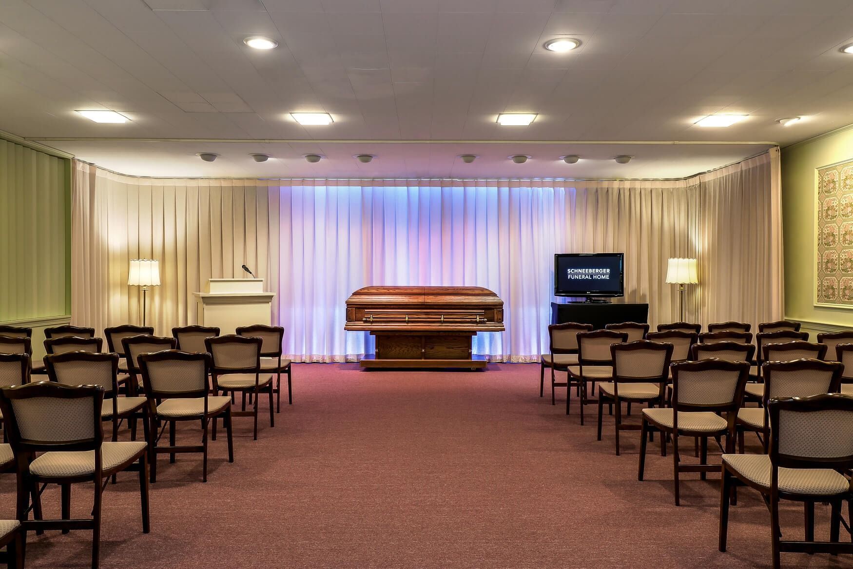 Empty funeral chapel with casket centered before a white draped backdrop and rows of chairs facing it