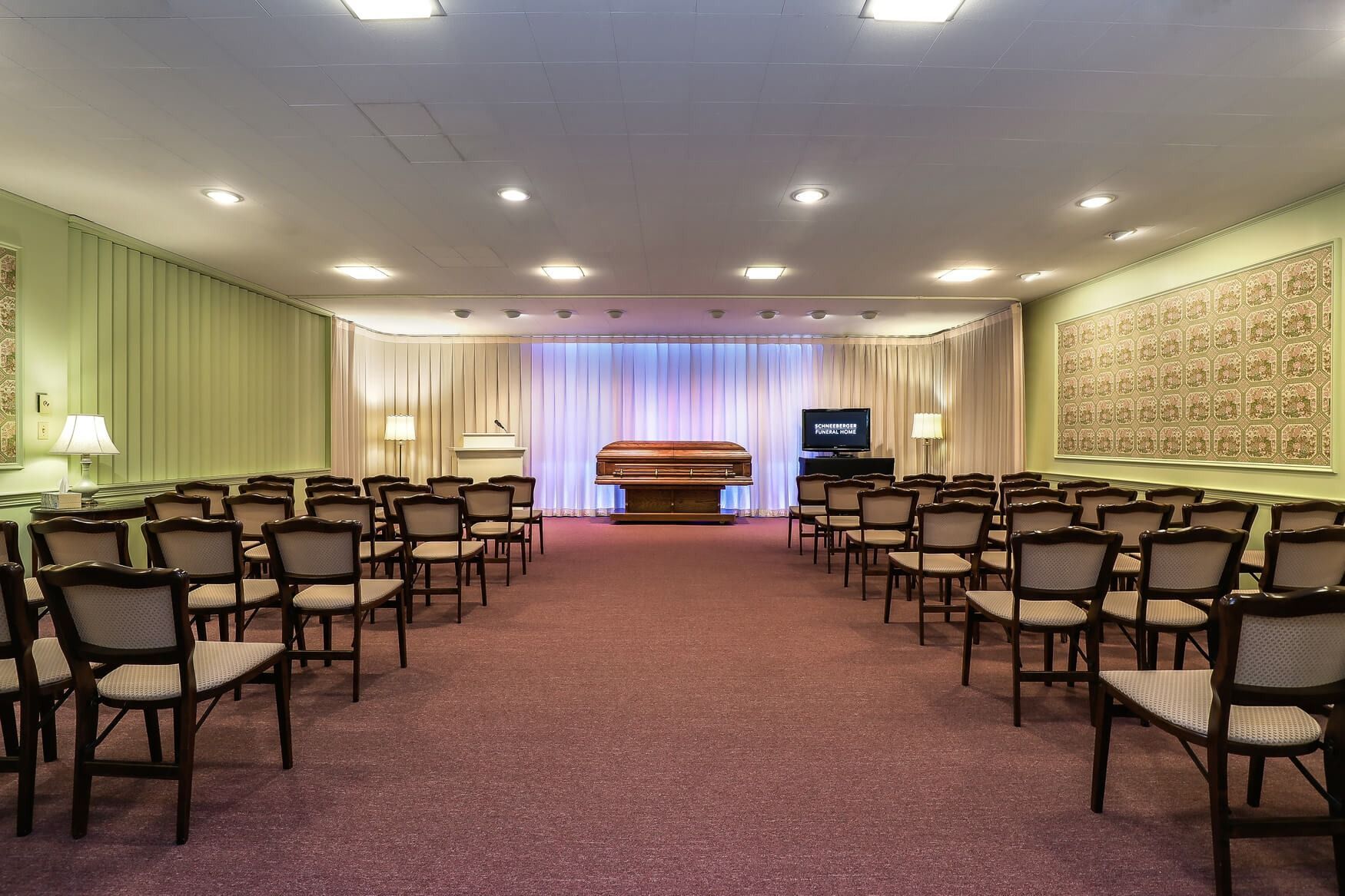 Empty chapel with rows of chairs facing a casket on a raised platform, lit by soft ceiling lights