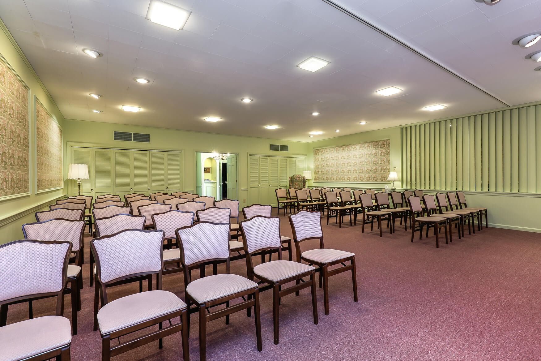Empty banquet hall with rows of chairs, pink carpet, and pale green walls under bright ceiling lights