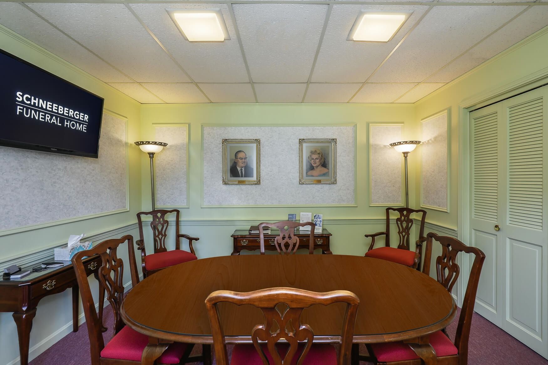 Small conference room with oval wooden table, red chairs, wall art, and lamps.