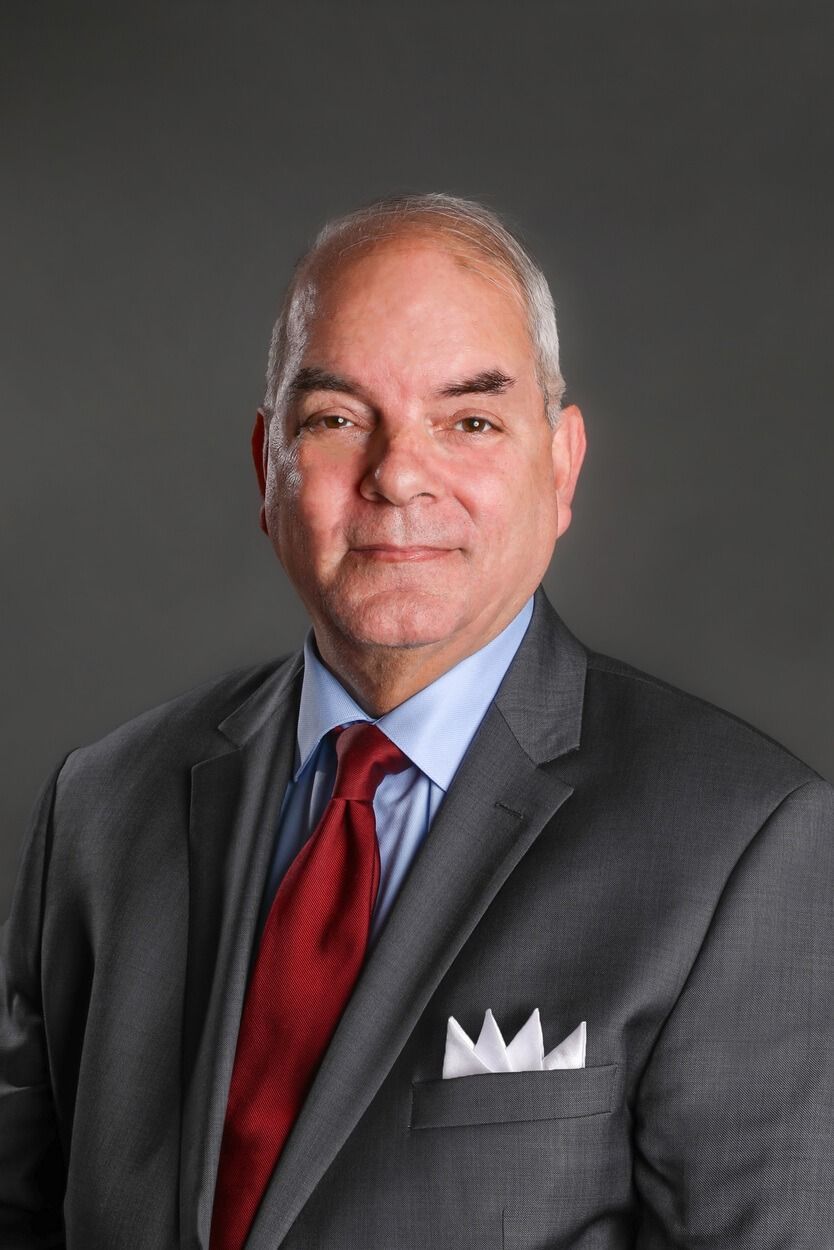 Studio portrait of a man in a gray suit, blue shirt, red tie, and white pocket square, against a dark background