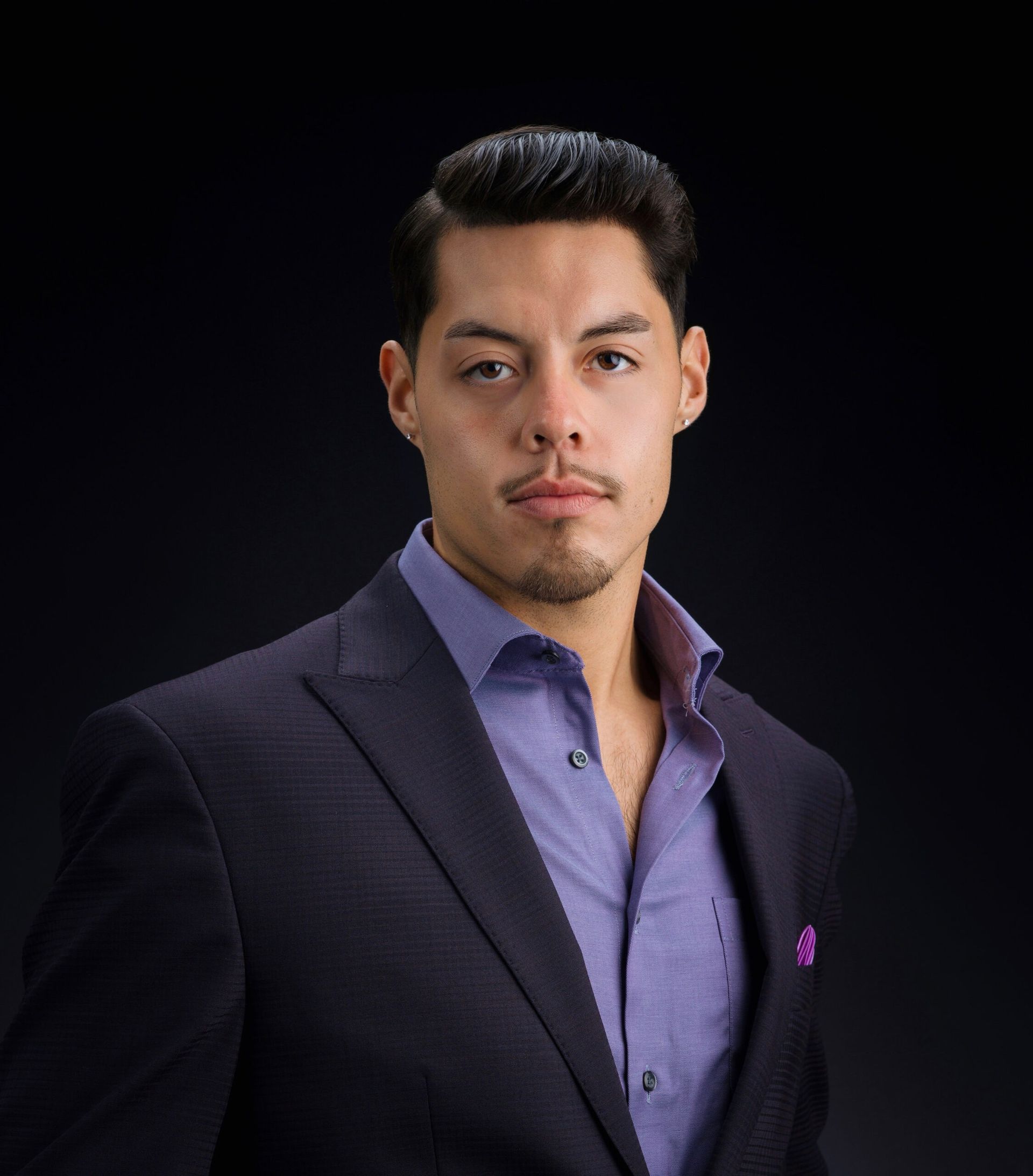 Man in a dark blazer and purple shirt posed against a black background.