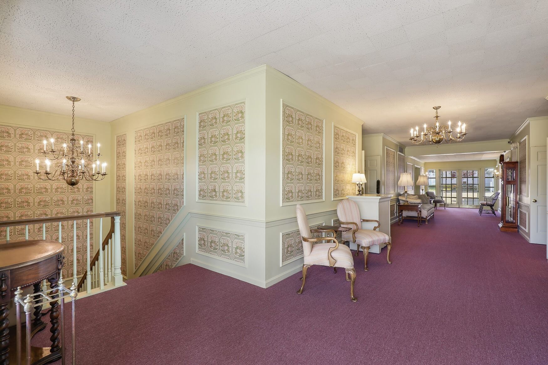 Elegant carpeted hallway with chandeliers and cream chairs overlooking a stairwell