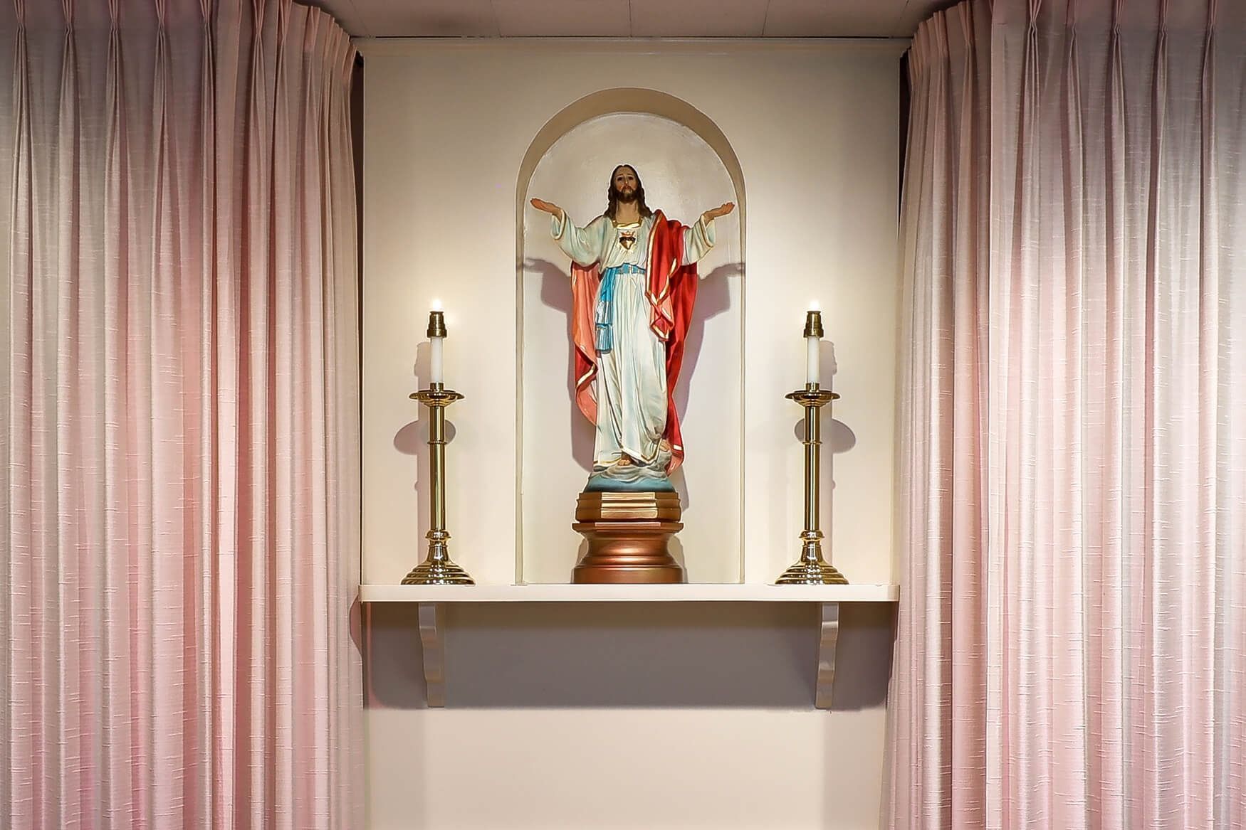 Small altar with a statue in red and blue robes, flanked by candles between pink curtains.