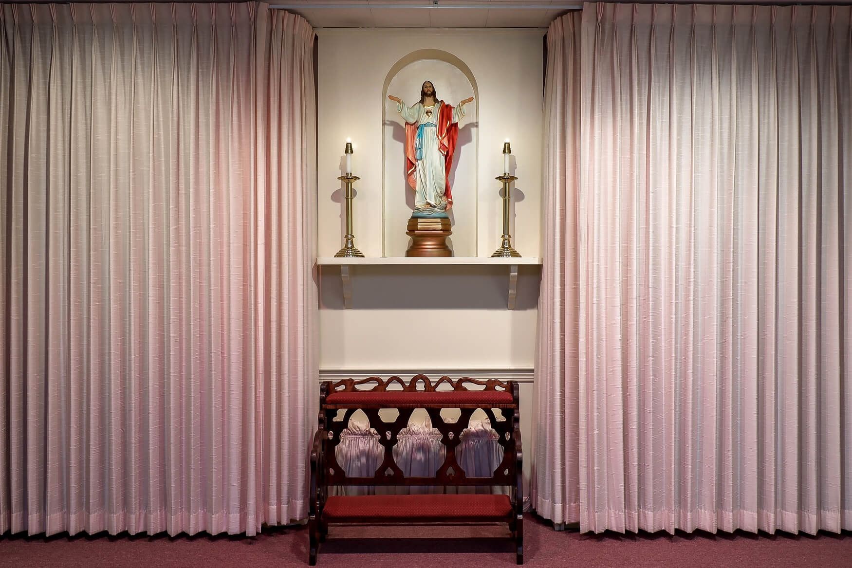 Small chapel altar with a red-robed statue, candles, and pink curtains behind a kneeler.