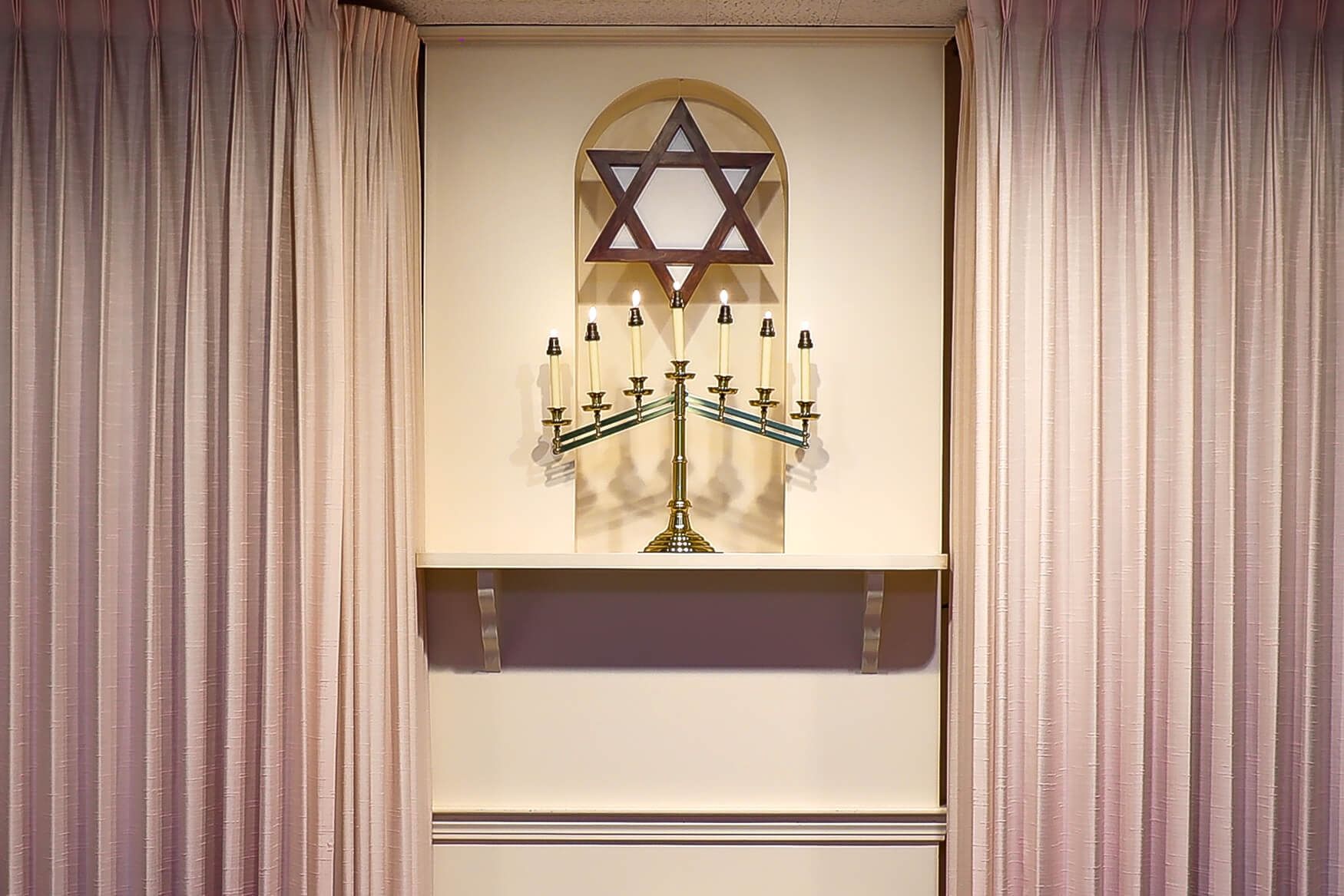 Star of David above a menorah on a wall niche, framed by closed pink curtains