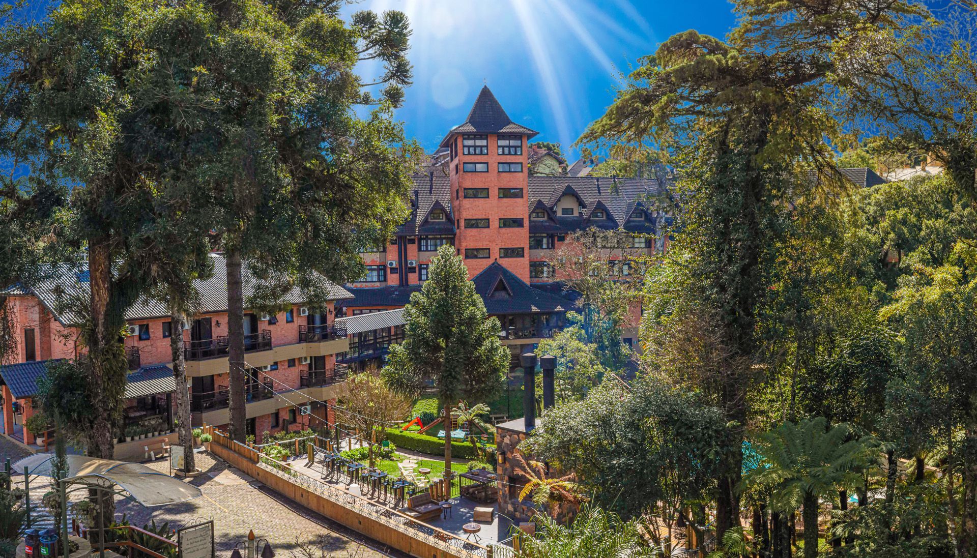 Parque ensolarado com árvores altas, uma fonte central e um edifício de tijolos vermelhos ao fundo.