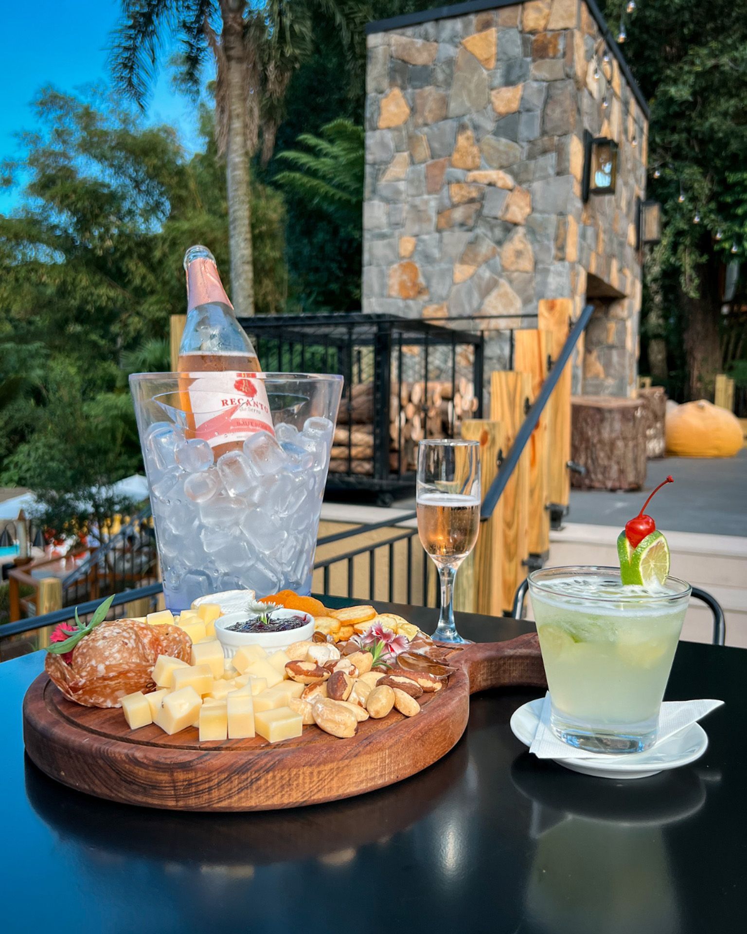 Mesa externa com tábua de queijos, balde de champanhe e dois coquetéis ao lado de um muro de pedra junto à piscina.