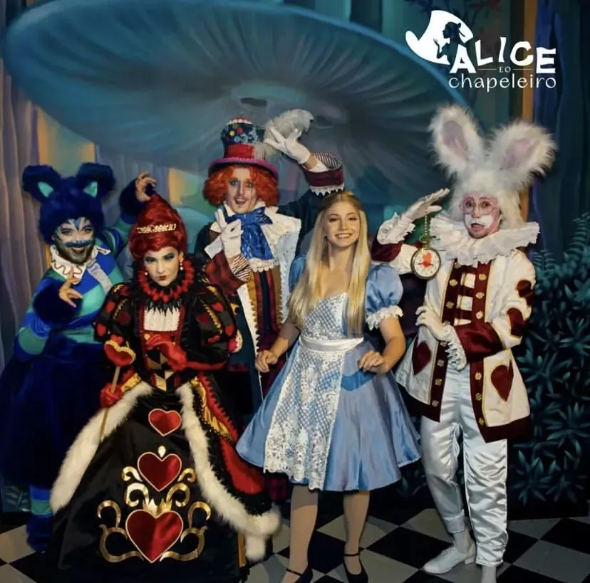 Grupo fantasiado posando em um cenário inspirado em Alice no País das Maravilhas, com um grande cogumelo azul ao fundo.