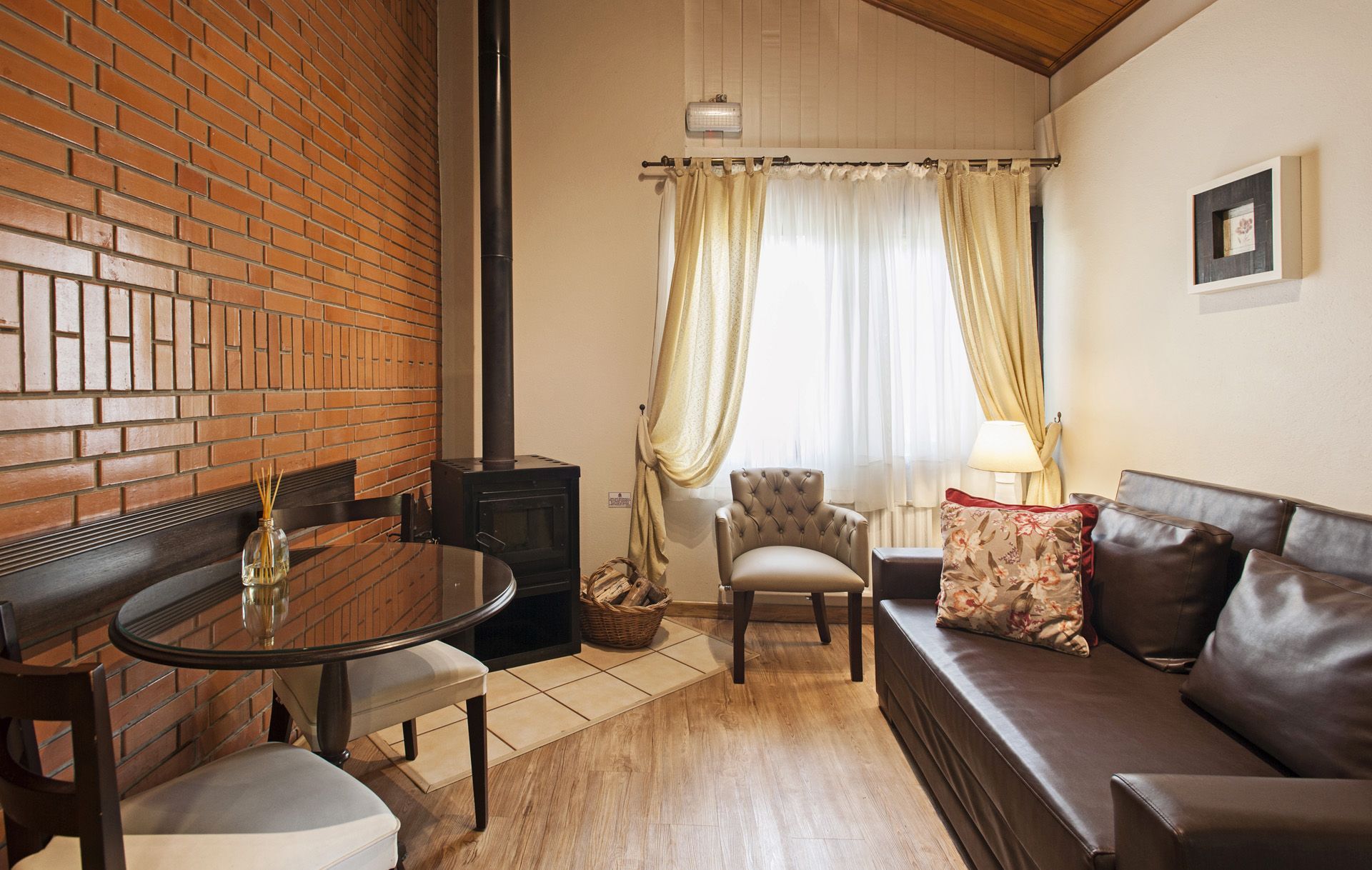 Sala de estar aconchegante com parede de tijolos, sofá de couro, mesa redonda e janela iluminada com cortinas.