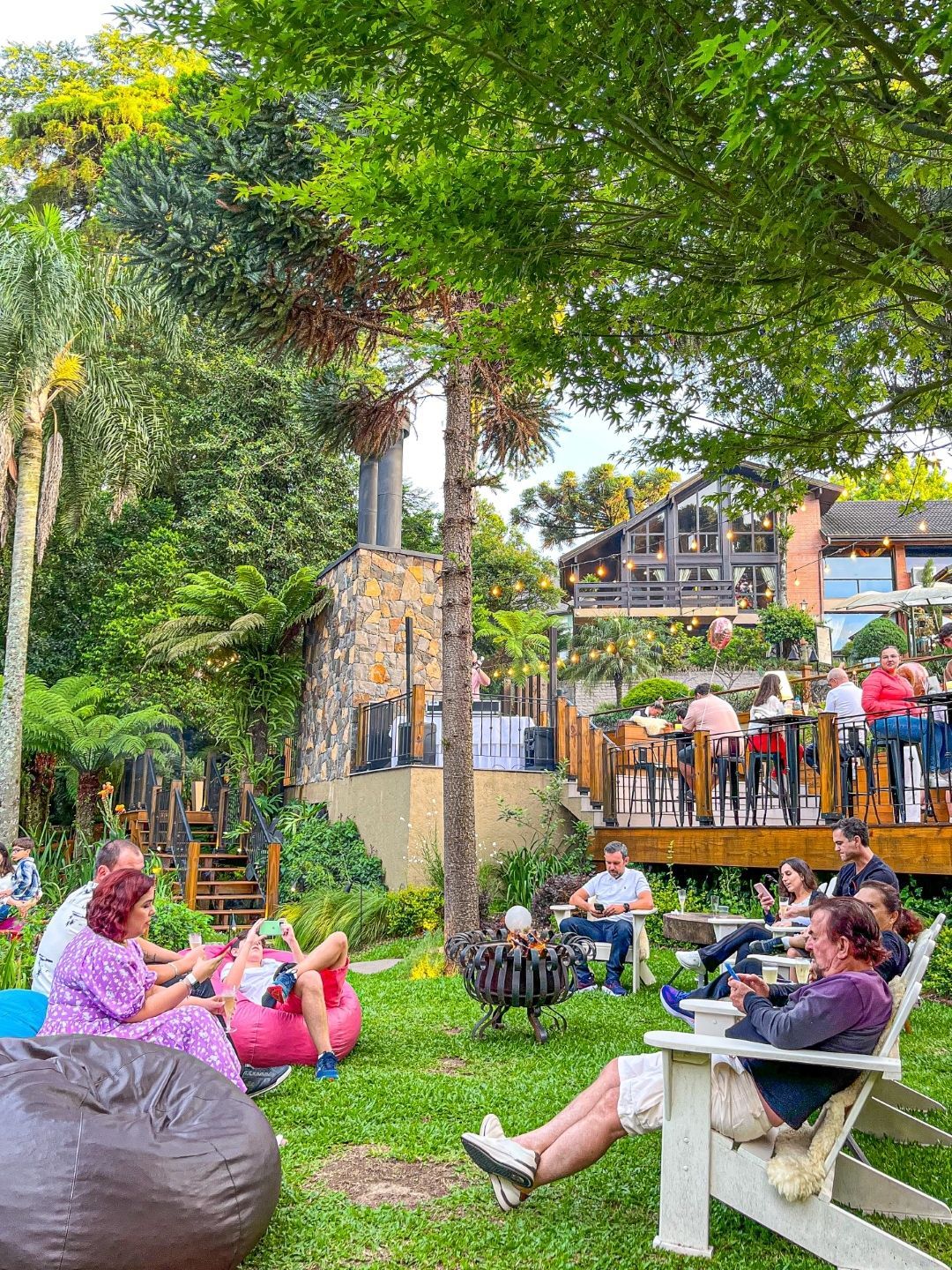 Pessoas relaxando em um parque verde com bancos, mantas de piquenique e um café com deck de madeira ao fundo.